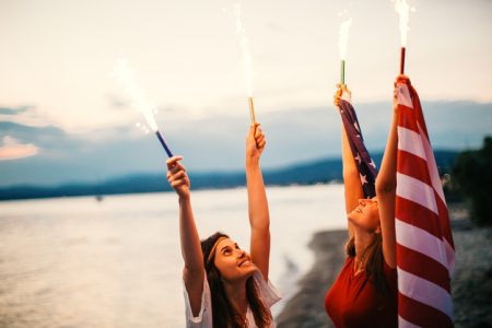 Young hipster girls at the beach, celebrating the fourth of July - by holding an American flag and fire torches up high