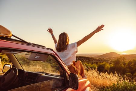 woman sitting on hood of car looking into the sunset with arms raised in the air